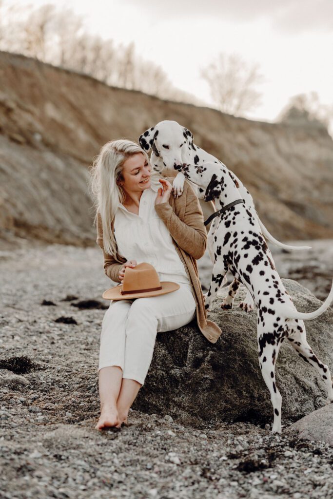 Frauchen Maxi sitzt mit ihren beiden Dalmatinern auf einem Felsen am Strand und spielt mit den Hunden.